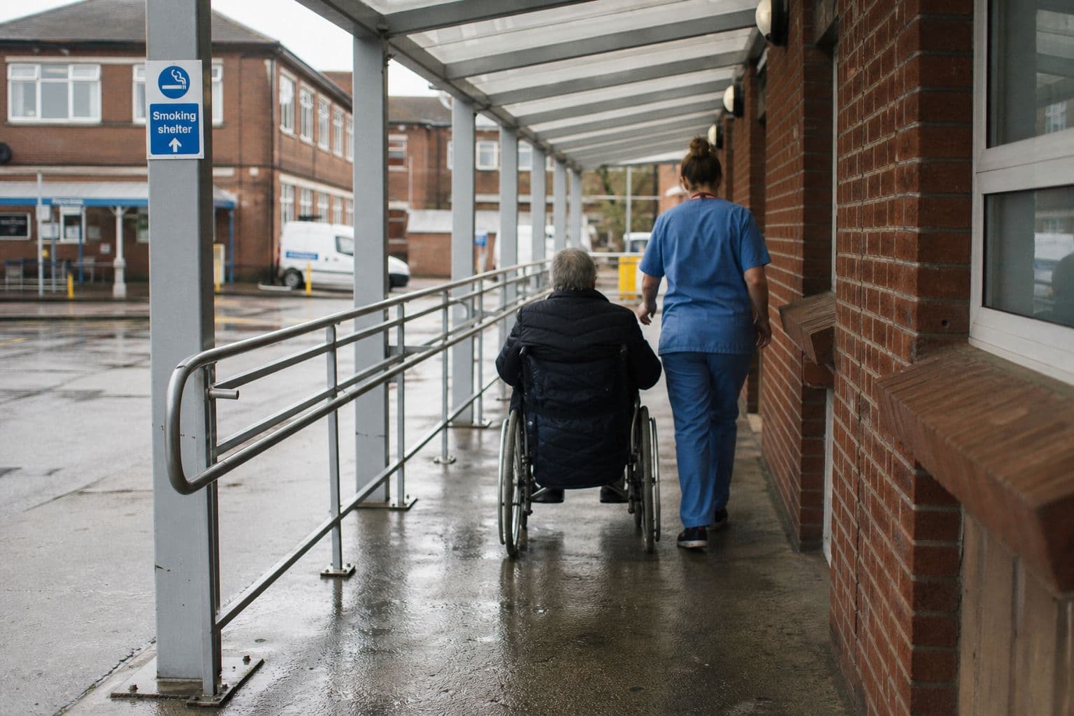Nurse and a wheelchair user under a covered walkway with handrails beside a hospital building