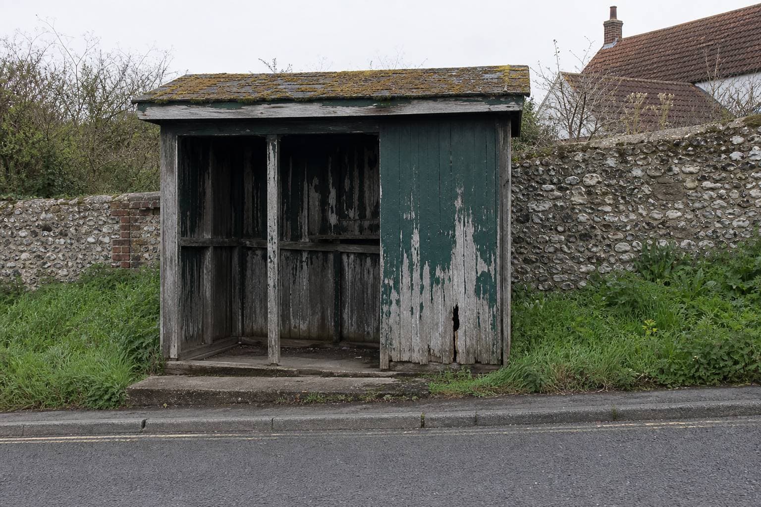 Weathered wooden bus shelter with peeling paint beside a flint wall