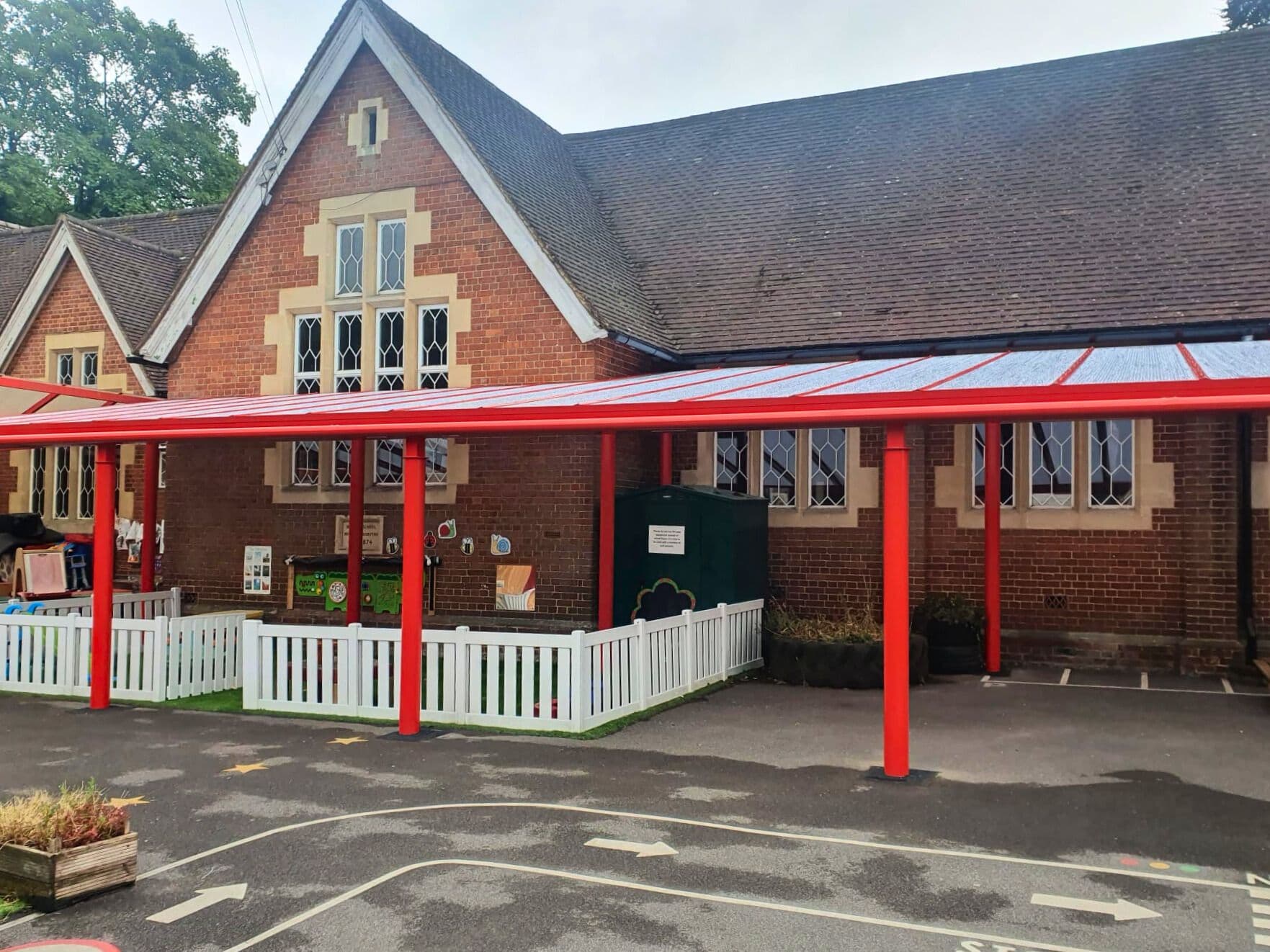 Canopy with red posts along a brick school building, covering a nursery play area