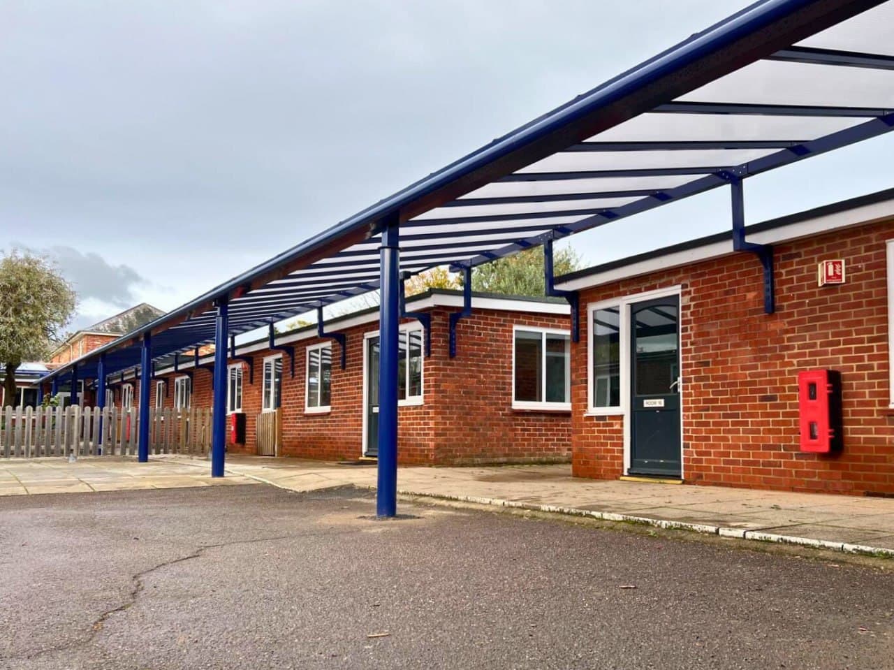 Blue covered walkway running the full length of a primary school building, long-run polycarbonate roof, aluminium posts