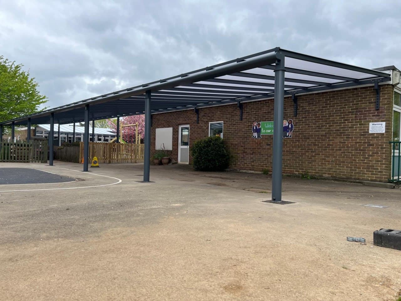 Large anthracite canopy covering school playground, flat polycarbonate roof, open-sided aluminium structure