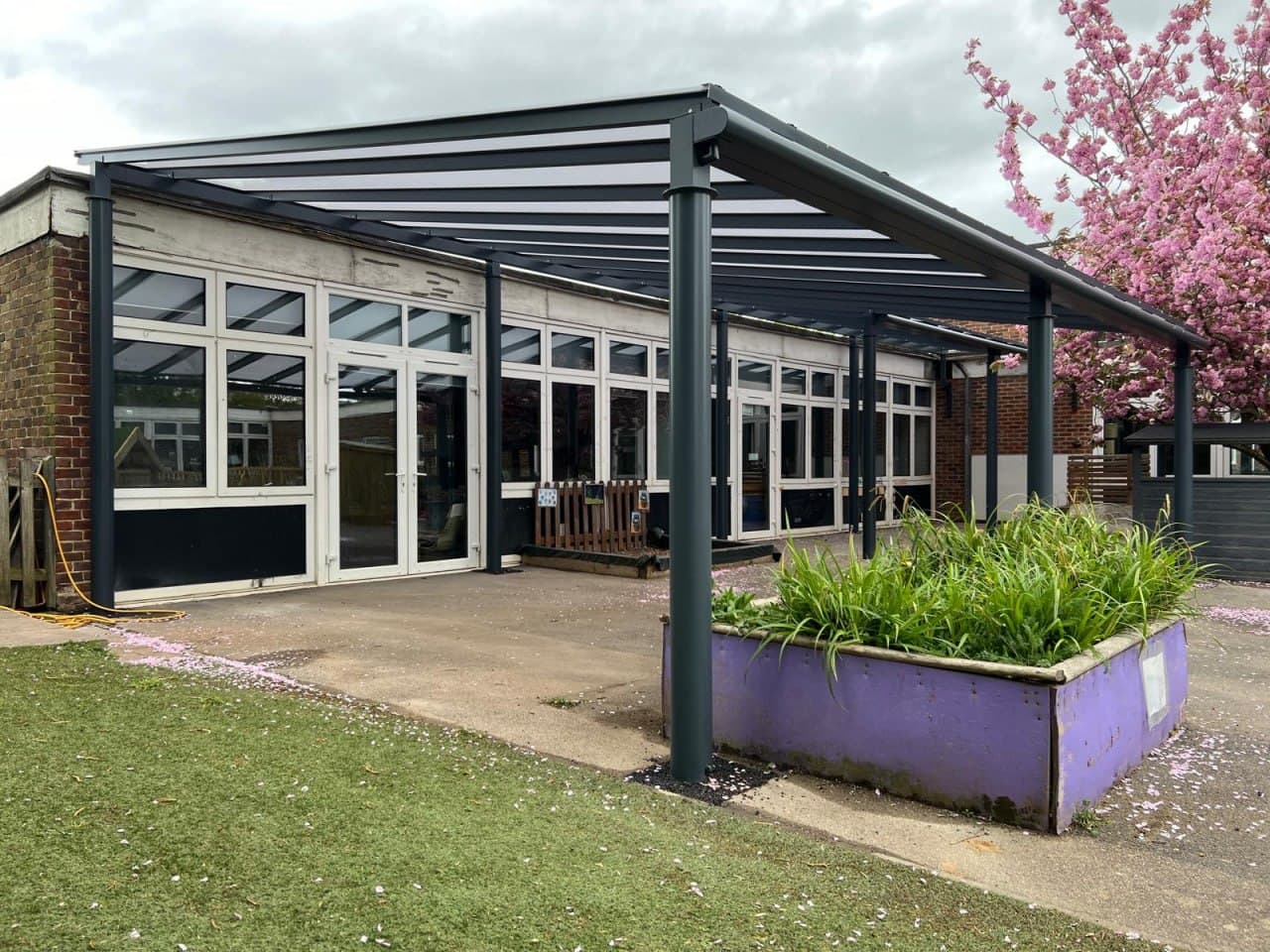Dark grey flat roof canopy at primary school with spring blossom trees, polycarbonate roofing on anthracite aluminium frame