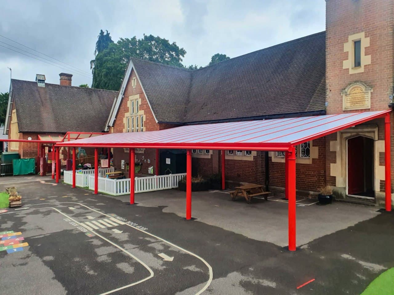 Red aluminium canopy covering primary school playground, polycarbonate flat roof, Victorian brick school building