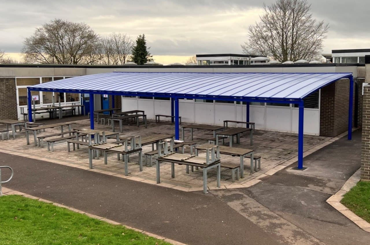 Large blue school dining canopy with flat polycarbonate roof covering outdoor picnic table area, wide-span aluminium structure