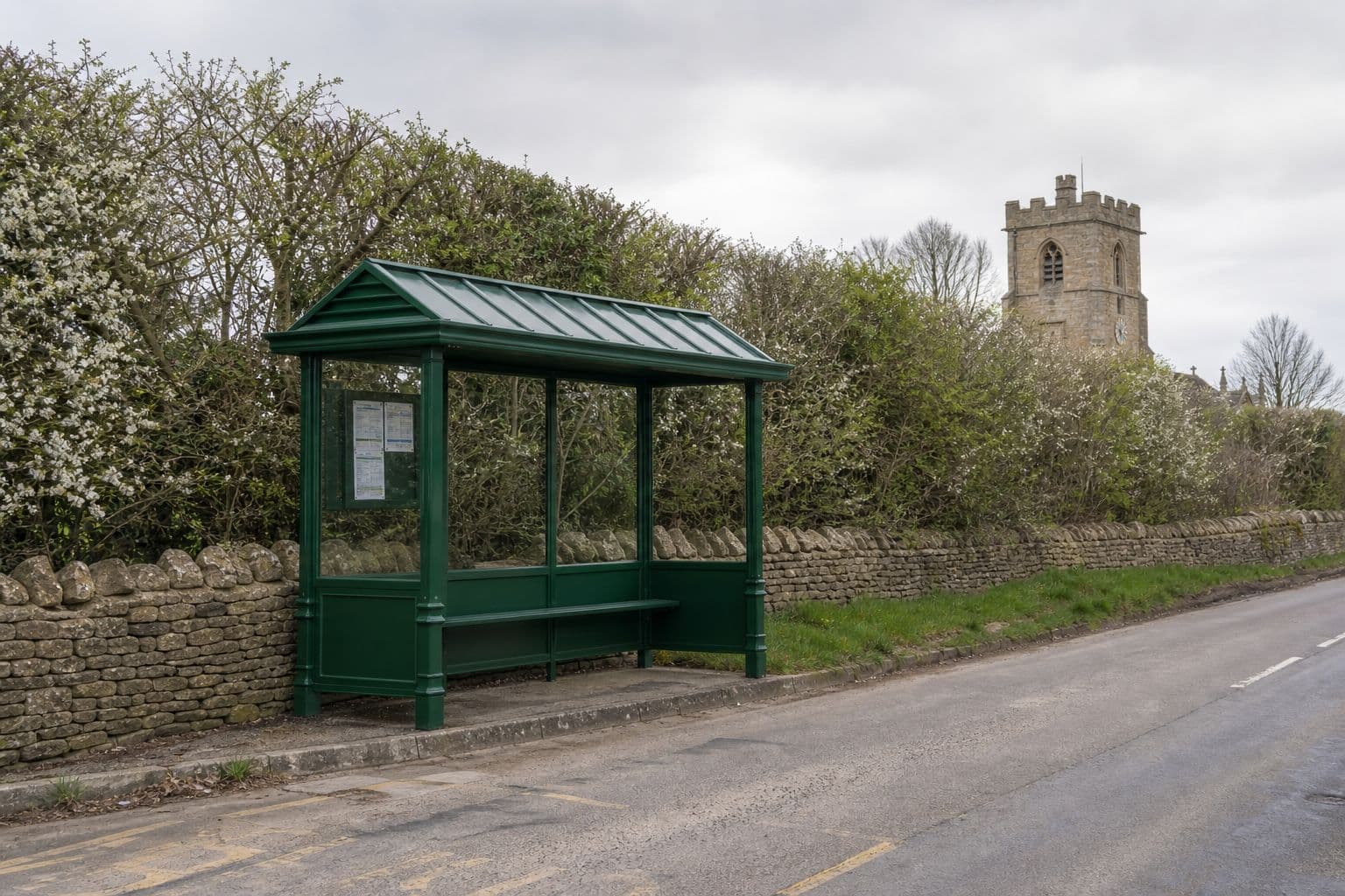 Green heritage-style bus shelter beside a dry stone wall on a village road, with a church tower behind