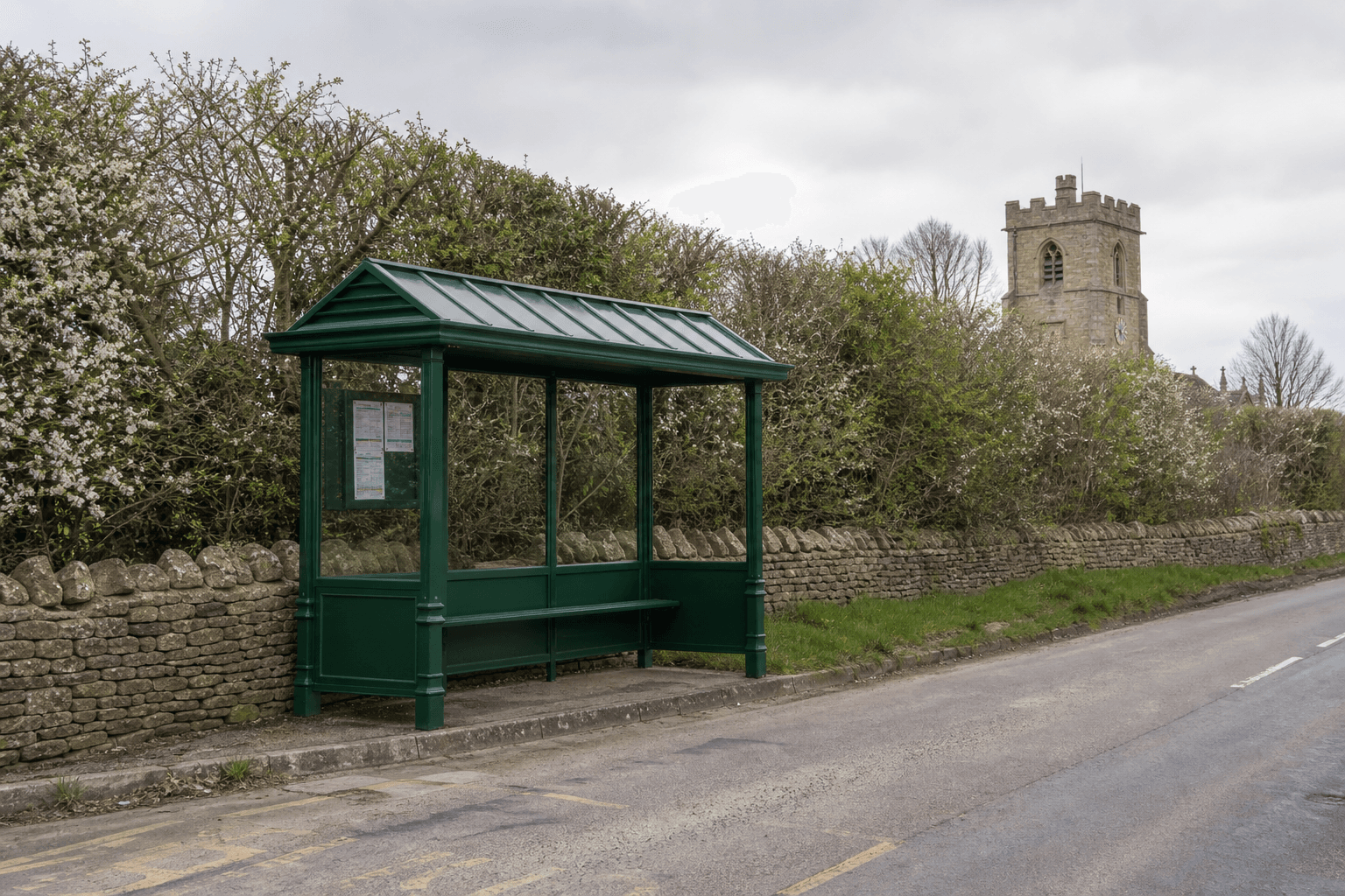Heritage-style green bus shelter in an English village beside a dry stone wall with a church tower visible in the background