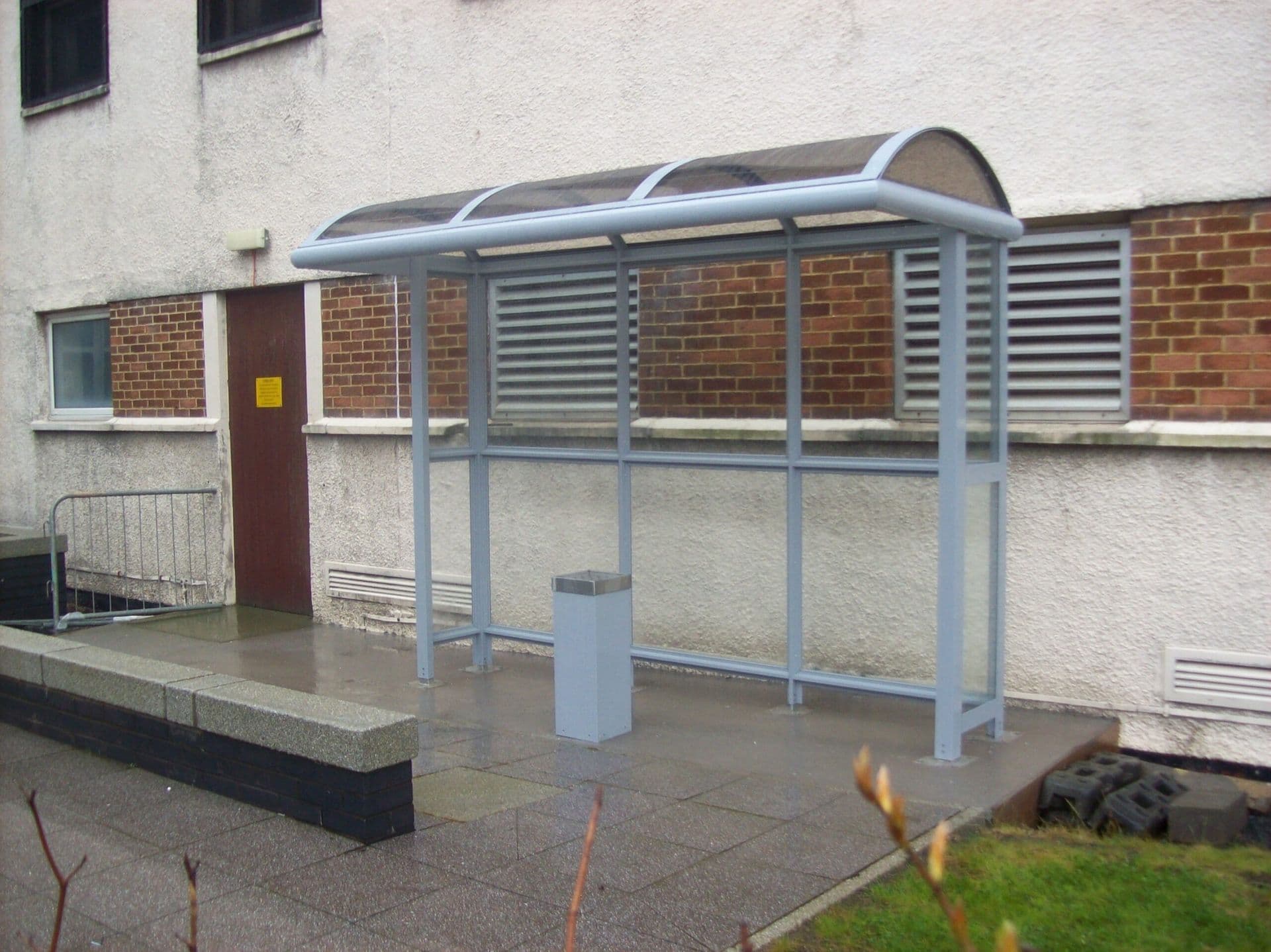 Grey smoking shelter with a curved clear roof against the wall of a commercial building
