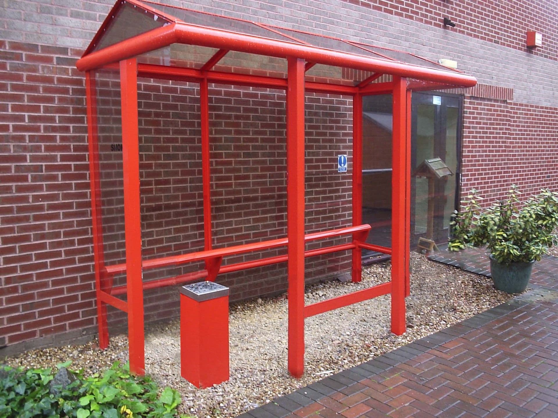 Red open-fronted smoking shelter with a cigarette bin beside an office building
